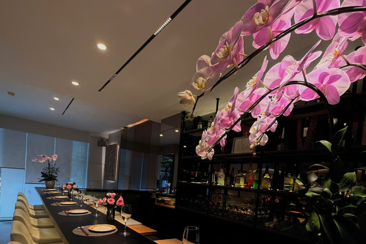 Large pink orchid blooms sweeping over the counter, with the lit sake and spirits shelf behind and place settings and seating visible along the black counter.