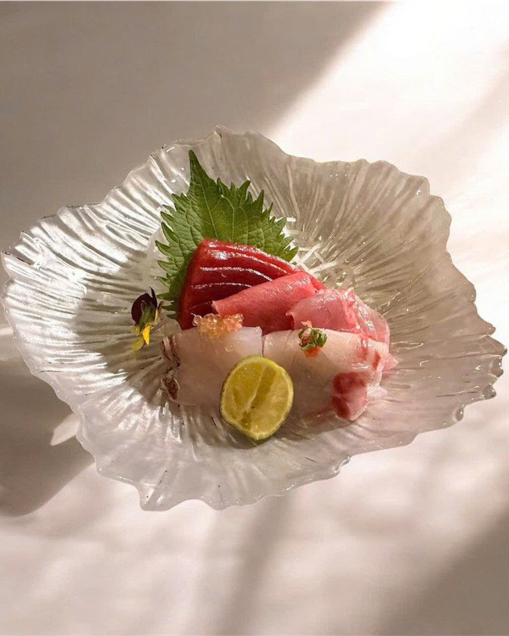 An assortment of sashimi with shiso leaf, edible flower and citrus, on a pale stoneware dish.