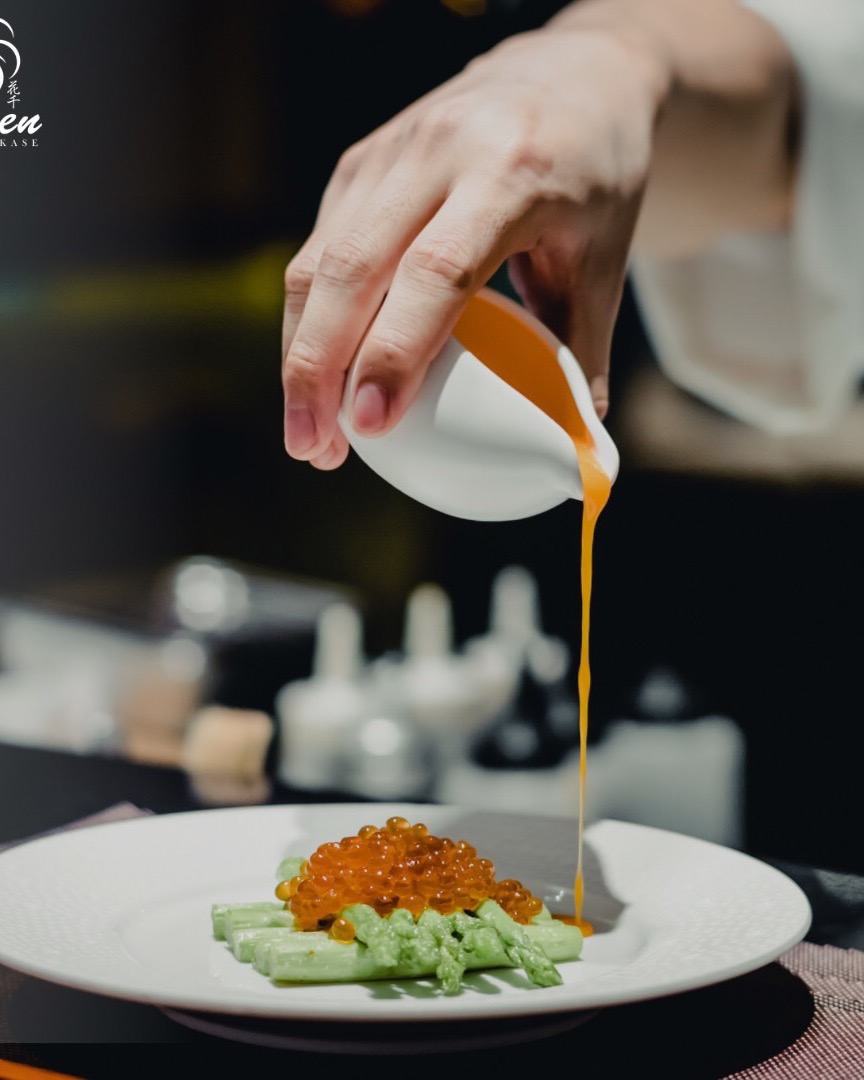 A chef's hand pouring sauce over a plate of ikura and asparagus.