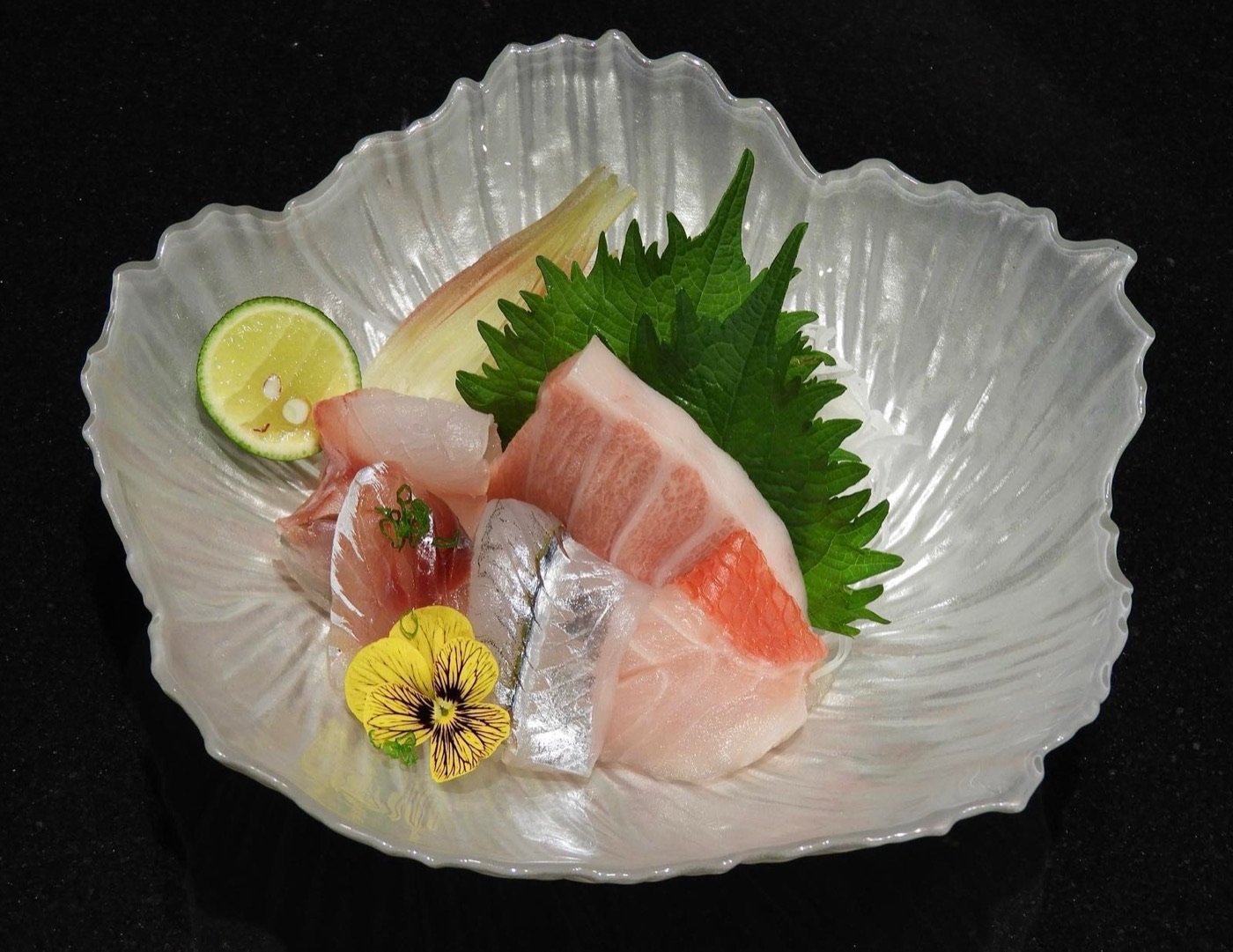 Một phần sashimi trên đĩa thủy tinh hình vỏ sò, cùng tía tô, cam chanh và một bông hoa ăn được.
