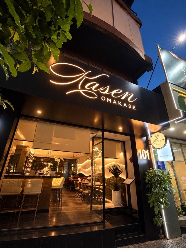 The illuminated Kasen Omakase sign above the entrance, framed by overhanging greenery.